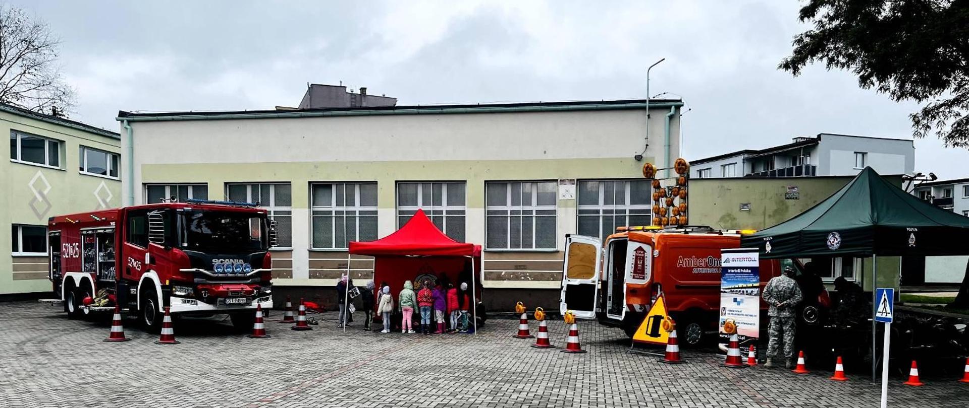 Zdjęcie przedstawia plac przed szkołą, służby prowadzą zajęcia edukacyjne, na pierwszym planie namiot i pojazdy PSP, w tle budynek szkoły