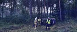 Przed leżącym na lewym boku autem trzech strażaków i dwóch policjantów. Dwóch strażaków na głowach ma czerwone, a trzeci biały hełm. Policjanci na głowach mają białe czapki. Jeden z nich, wyjmujący coś z kieszeni ma na sobie żółtą kamizelkę odblaskową. W tle - las.