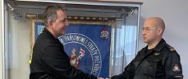 Dwóch mężczyzn w mundurach Państwowej Straży Pożarnej stoi naprzeciw siebie podając sobie ręce, mężczyzna z prawej strony trzyma w ręku dokument, za nimi widoczna jest gablota w której wisi sztandar Komendy powiatowej PSP w Pleszewie