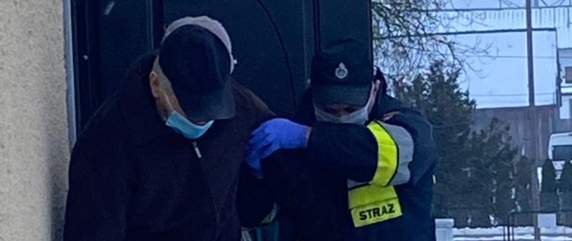 W centrum widać strażaka ubranego w ubranie specjalne, rękawiczki lateksowe i maseczkę. Strażak pomaga osobie starszej, poruszającej się o lasce zejść ze schodów.