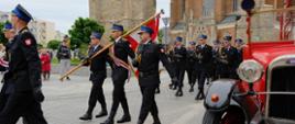 Uroczysty przemarsz pododdziału strażaków w galowych mundurach przed kościołem. Na czele kolumny niesiona jest flaga państwowa. Strażacy kroczą w równym szyku, w tle widoczne są gotyckie mury kościoła i wieża. Po prawej stronie kadru stoi zabytkowy, czerwony samochód pożarniczy.