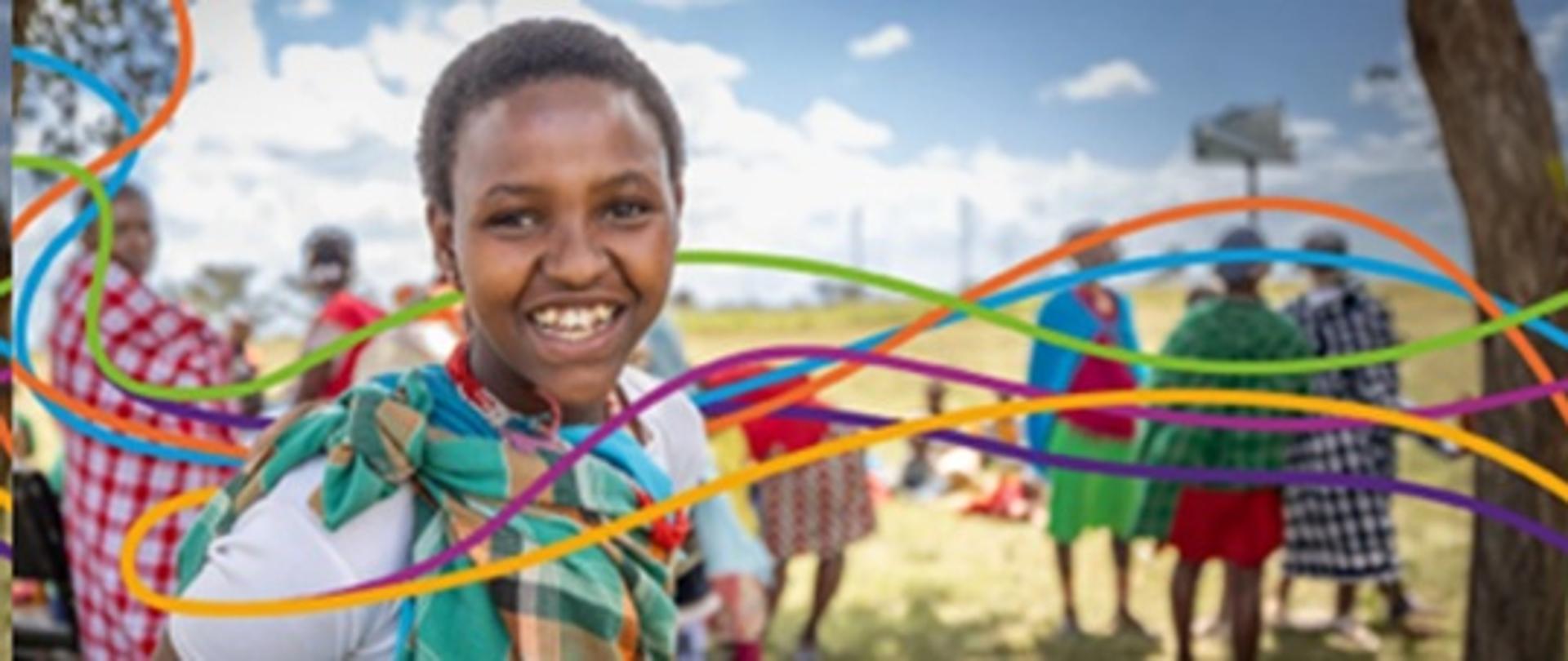 na zdjęciu dziecko rasy czarnej z krótkimi włosami uśmiecha się w tle stoją kolorowo ubrani ludzie