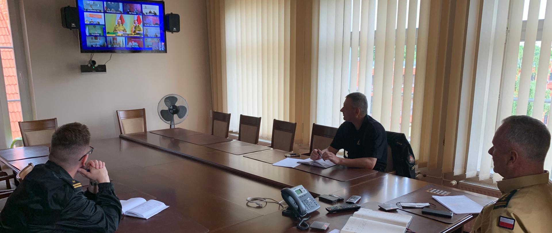 Na zdjęciu widać trzech funkcjonariuszy siedzących przy konferencyjnym stole i patrzących w ekran telewizora, na którym odbywa się wideokonferencja komendantów wojewódzkich PSP z komendantem głównym PSP.