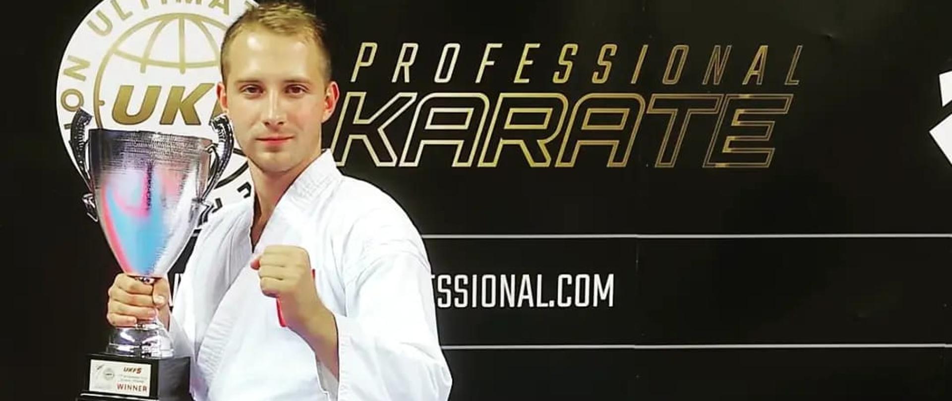 Na zdjęciu widać mężczyznę, zawodnika karate w białym stroju z czarnym pasem. W ręku trzyma puchar. W tle jest czarna plansza na której są częściowo zasłonięte napisy. Jeden z napisów na planszy to professional karate.
