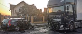 Na zdjęciu dwa rozbite pojazdy osobowy i ciężarowy po zderzeniu czołowym, znacznie zdeformowane na drodze asfaltowej. W tle budynek mieszkalny i zachodzące słońce.