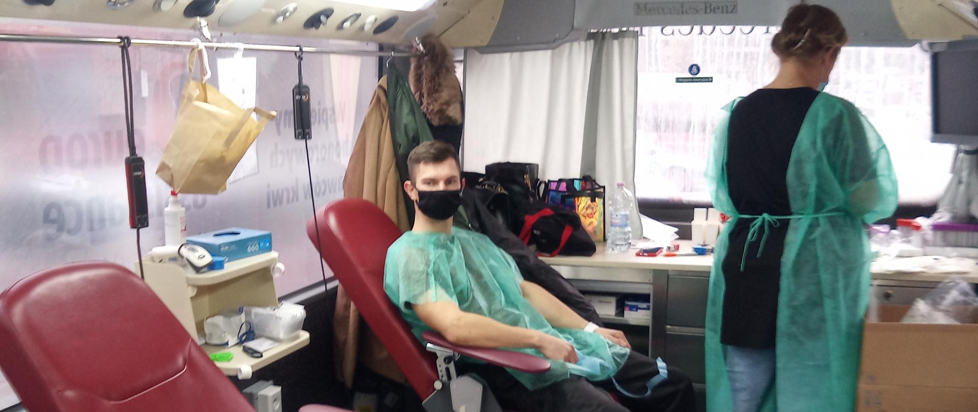 Mężczyzna siedzący na fotelu z jedną ręką przygotowaną do oddawania krwi. Obok niego stoi kobieta.