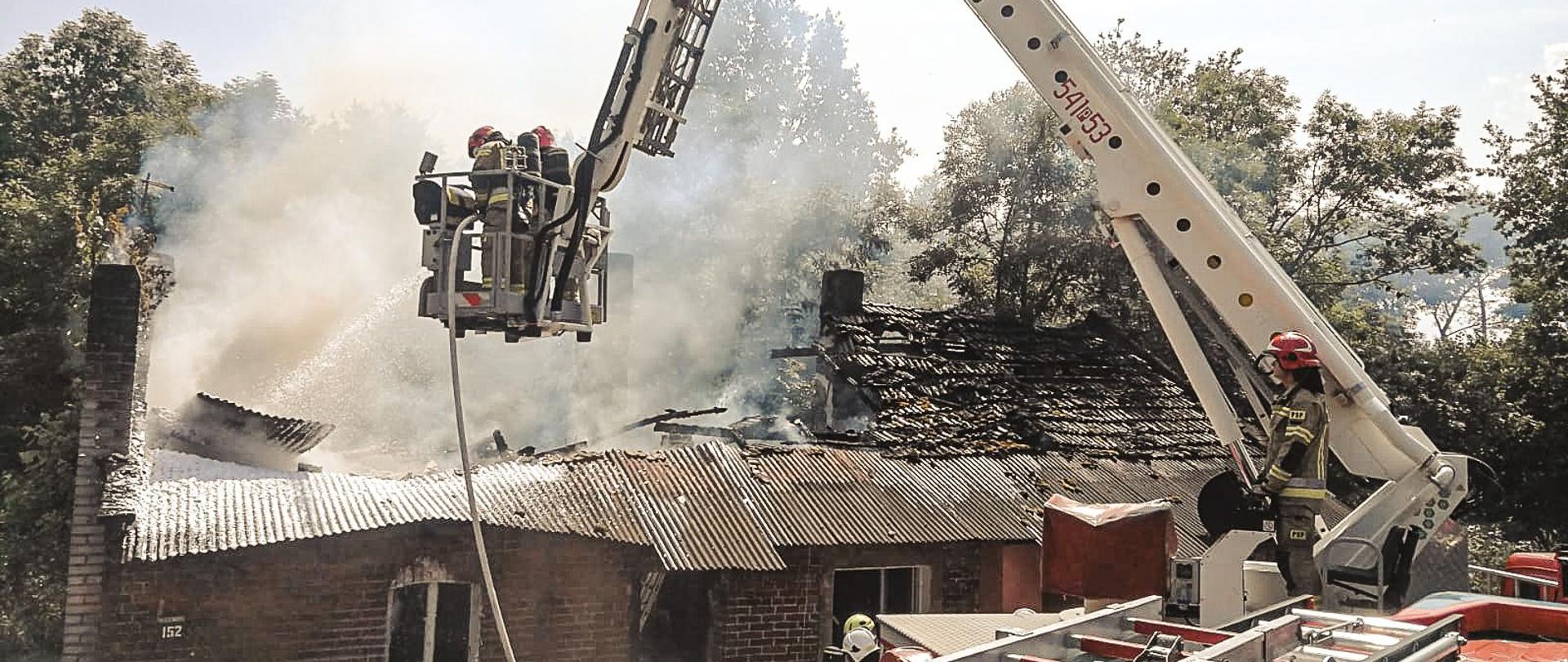 Strażacy stojący przy samochodach strażackich. Na ziemi leżą węże pożarnicze. Na drugim planie palący się obiekt.