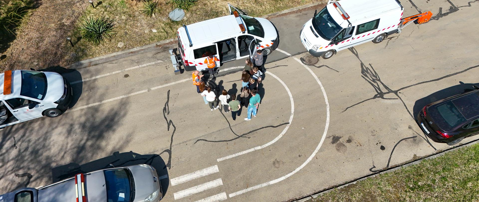 Na zdjęciu grupa osób stoi wokół białego samochodu dostawczego z pomarańczowo-białymi pasami. Drzwi pojazdu są otwarte, a dwie osoby w pomarańczowych kamizelkach odblaskowych prowadzą aktywną interakcję z grupą. W pobliżu zaparkowane są inne pojazdy, w tym drugi biały samochód dostawczy o podobnych oznaczeniach oraz srebrny samochód osobowy. Scena rozgrywa się na utwardzonym terenie z oznakowanymi pasami i przejściem dla pieszych. Otoczenie i detale sugerują, że jest to terenowe szkolenie lub demonstracja praktyczna związana z budownictwem drogowym, gdzie uczestnicy mają okazję zdobywać wiedzę w rzeczywistych warunkach pracy