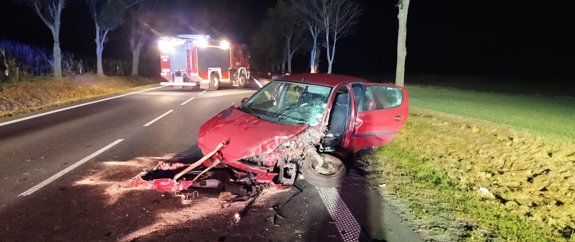 Na zdjęciu widać wrak rozbitego pojazdu osobowego, czerwony. Pojazd stoi częściowo na drodze, częściowo na poboczu. Ma rozbity cały przód. W tle samochód strażacki oświetlający teren nocą.