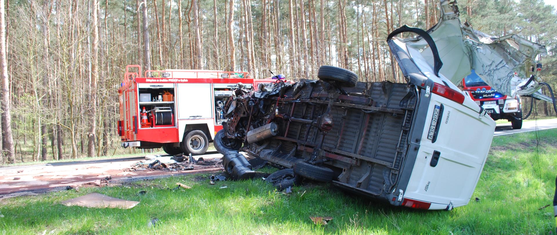 Zdjęcie przedstawia przewrócony na prawy bok wrak pojazdu dostawczego znajdujący się częściowo na poboczu drogi, w tle na jezdni widać dwa pojazdy pożarnicze