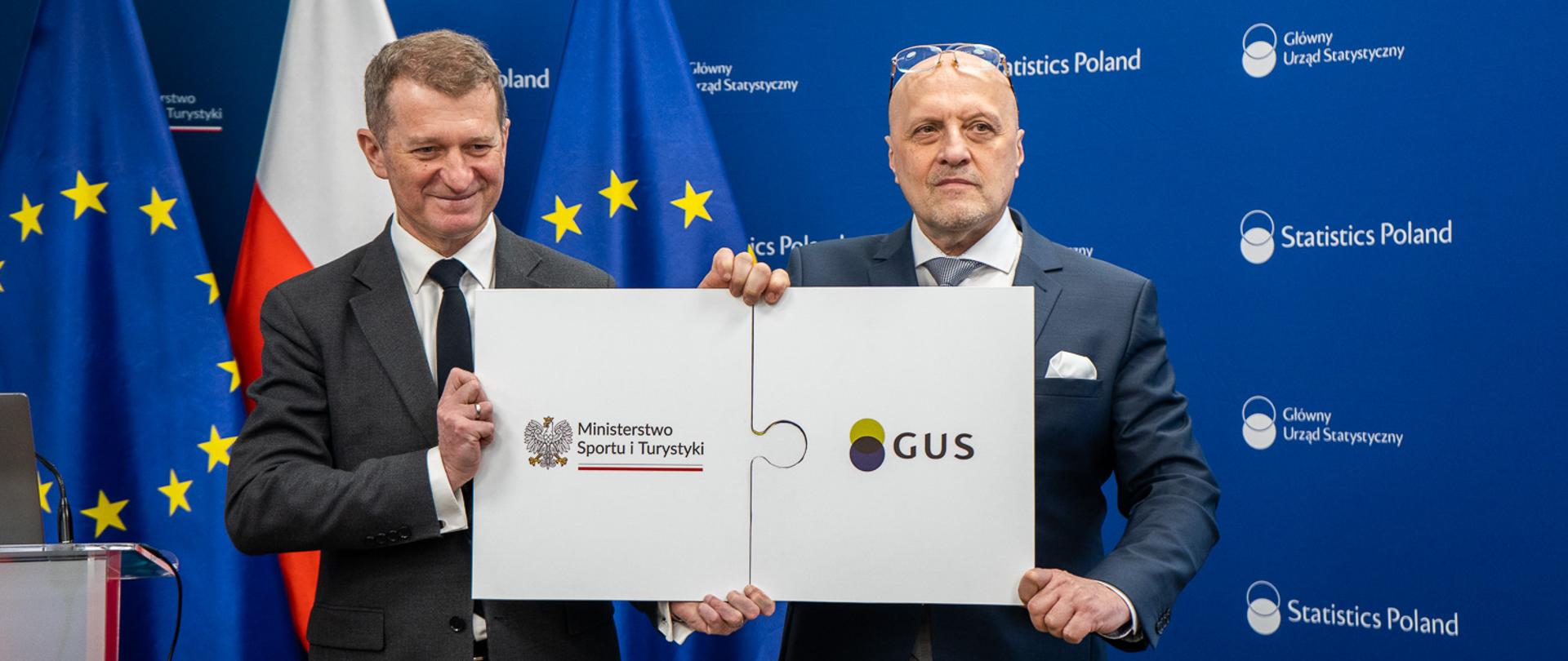 Wiceminister Ireneusz Raś z prezesem GUS Markiem Cierpiałem-Wolanem podczas konferencji nt. Obserwatorium Turystycznego. Stoją na tle flagi Polski oraz Unii Europejskiej. Trzymają w ręku plansze w kształcie dwóch puzzli z logotypem ministerstwa oraz GUS