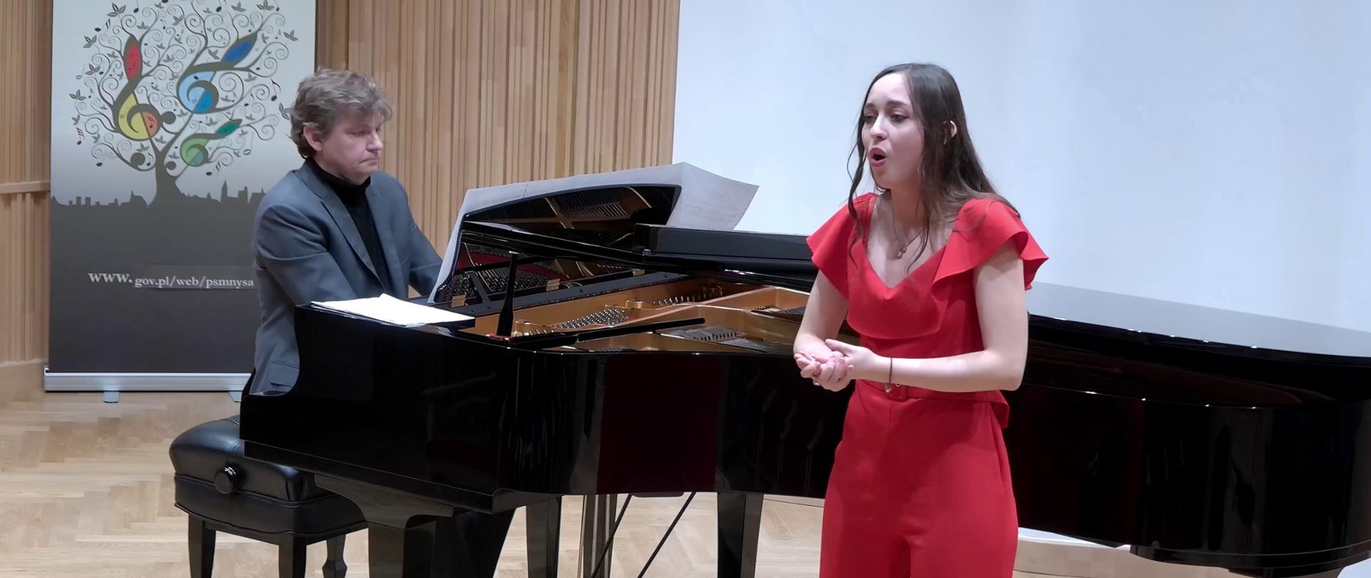 Zdjęcie. Na estradzie występuje ubrana w czerwony strój śpiewaczka z towarzyszeniem pianisty, który gra na dużym czarnym fortepianie. W głębi z lewej strony widać baner PSM Nysa.