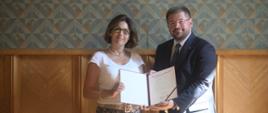 Wiceminister Rzymkowski stoi obok kobiety w białej bluzce i spódnicy w kwiatki, razem trzymają otwartą teczkę z dokumentem.