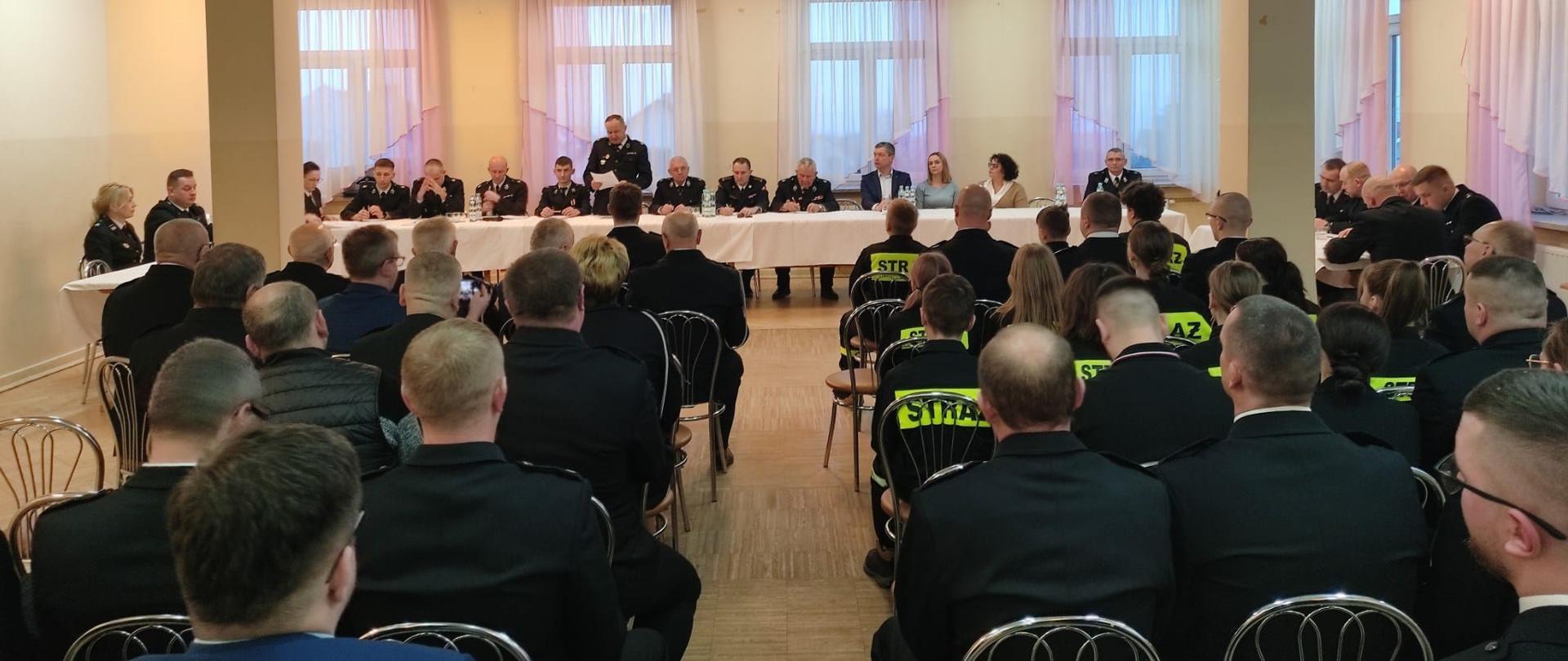 Zdjęcie przedstawia w głębi stół prezydialny gdzie siedzą zaproszeni goście wśród nich Zastępca Komendanta Miejskiego PSP w Kielcach st. bryg. Marcin Charuba i organizatorzy oraz przybyłych na spotkanie druhów – bliżej obserwującego. 