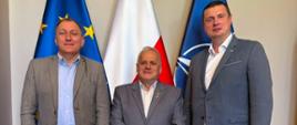 3 mężczyzn w garniturach, w tym wicewojewoda, pozują na tle 3 flag: UE, Polski i Nato
