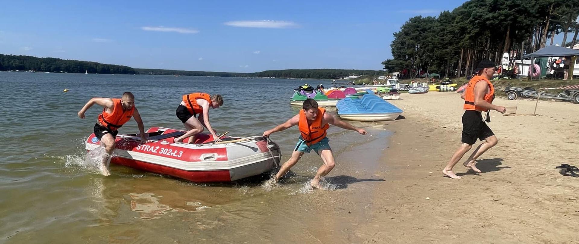 Na zdjęciu widzimy ratowników ubranych w kamizeli ratunkowe wyskakujących z łodzi na brzeg po wykonanej konkurencji. W tle piękna sceneria zbiornika wodnego i rowerków wodnych.