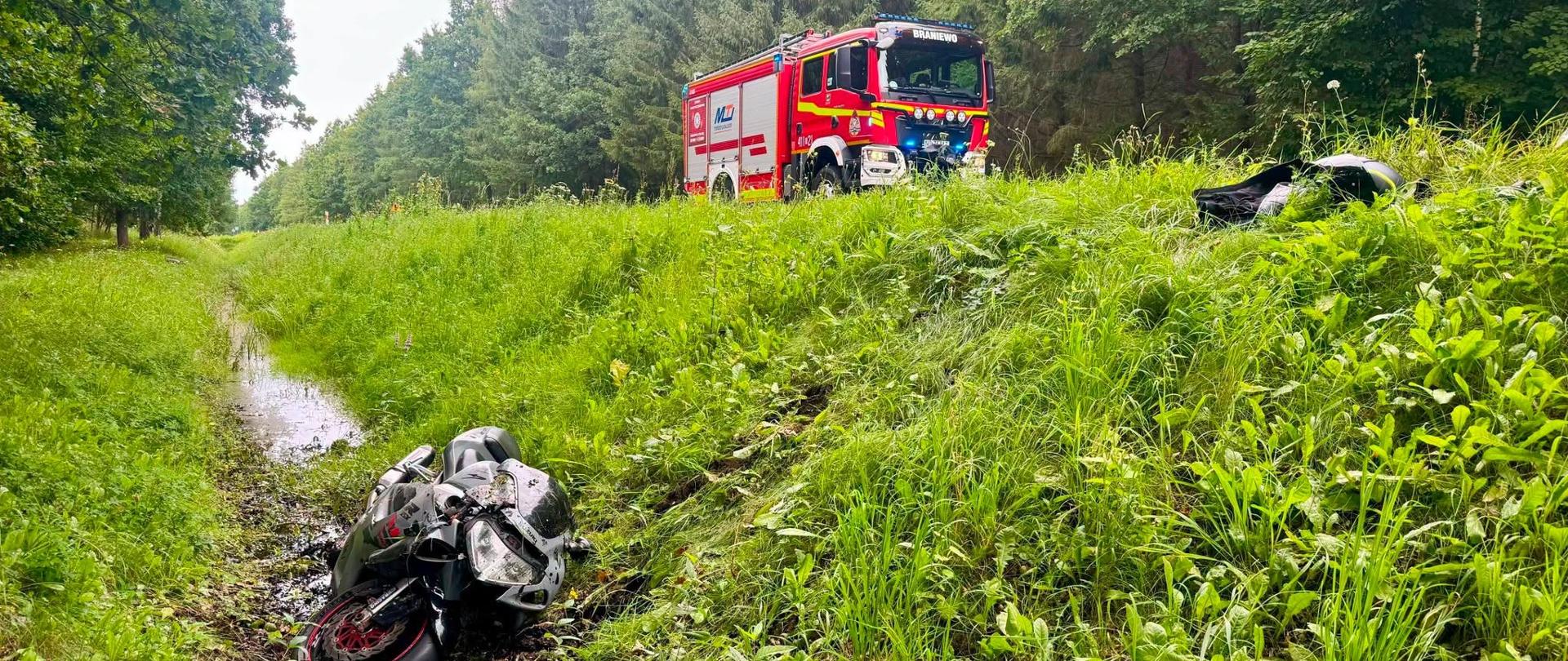 Na zdjęciu widać miejsce zdarzenia drogowego w lesie. W przydrożnym, podmokłym rowie leży przewrócony motocykl, a na poboczu drogi stoi czerwony samochód ratowniczo-gaśniczy Państwowej Straży Pożarnej z włączonymi światłami ostrzegawczymi. Teren wokół porośnięty jest wysoką trawą i gęstymi drzewami
