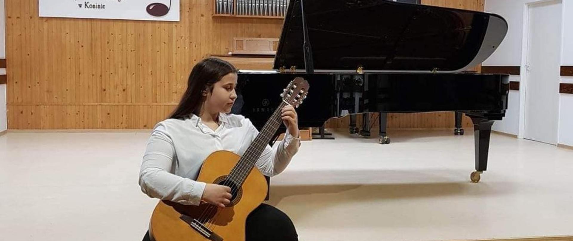 Zdjęcie przedstawia Patrycję w stroju galowym jak siedzi na scenie i gra na gitarze, w tle fortepian i portret Paderewskiego.