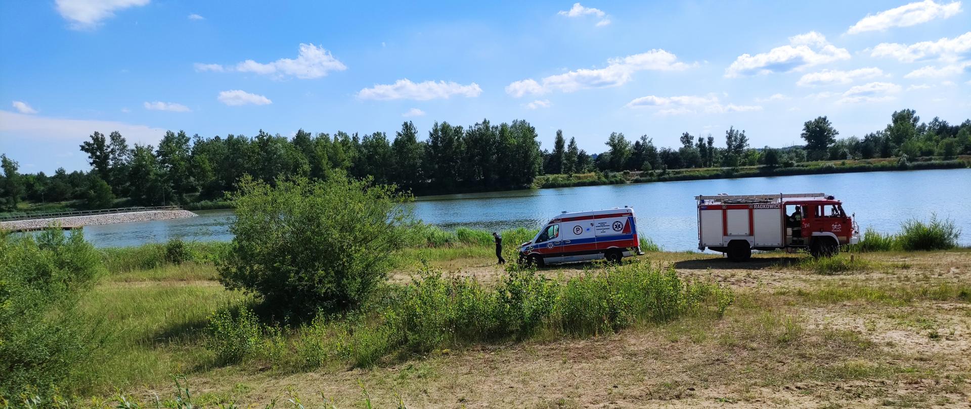 Zdjęcie przedstawia zbiornik wodny oraz przyległy do niego teren na którym stoi samochód strażacki oraz karetka pogotowia.