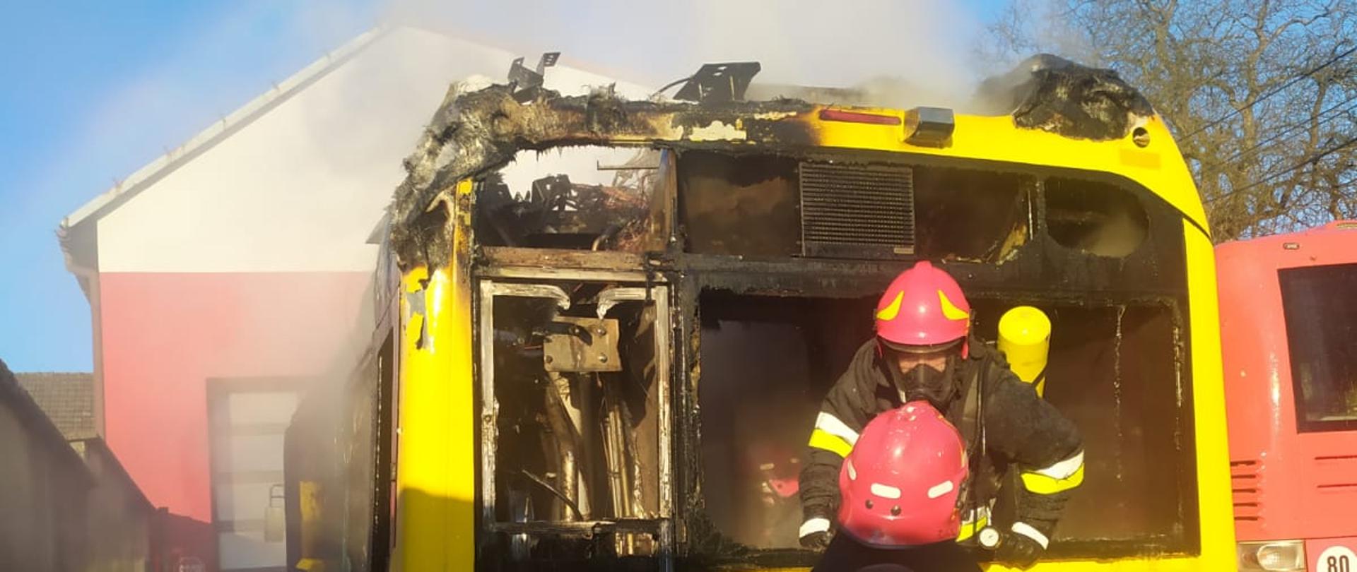 Na zdjęciu widoczny jest tył autobusu w kolorze żółtym. Strażacy w zabezpieczeniu prowadzą działania gaśnicze wewnątrz pojazdu. W górnej części widoczne są spalone elementy oraz wydobywający się z wnętrza dym.