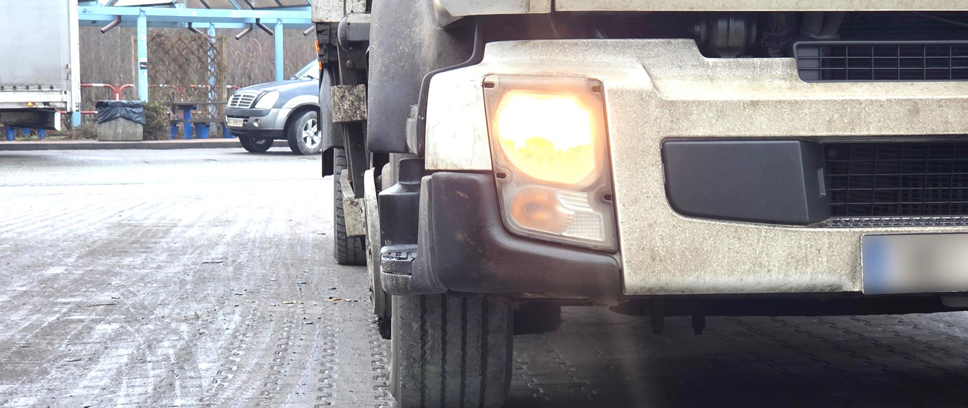 Zdjęcie przedstawia zbliżenie na przód ciężarówki zaparkowanej na wybrukowanym placu. Widoczny jest jedynie prawy dolny róg białej ciężarówki z włączonym żołtawym światłem. W tle po lewej stronie zdjęcia widoczny jest fragment samochodu osobowego oraz infrastruktura w postaci wiaty z ławeczkami i stołem, biało-czerwonymi barierkami, koszem na śmieci. Daleko w tle widoczny jest również las. W lewym górnym rogu zdjęcia, widoczny jest również fragment plandeki pojazdu. Nawierzchnia, na której stoi pojazd ciężarowy jest przybrudzona. Sceneria wskazuje na porę jesienną lub zimową.