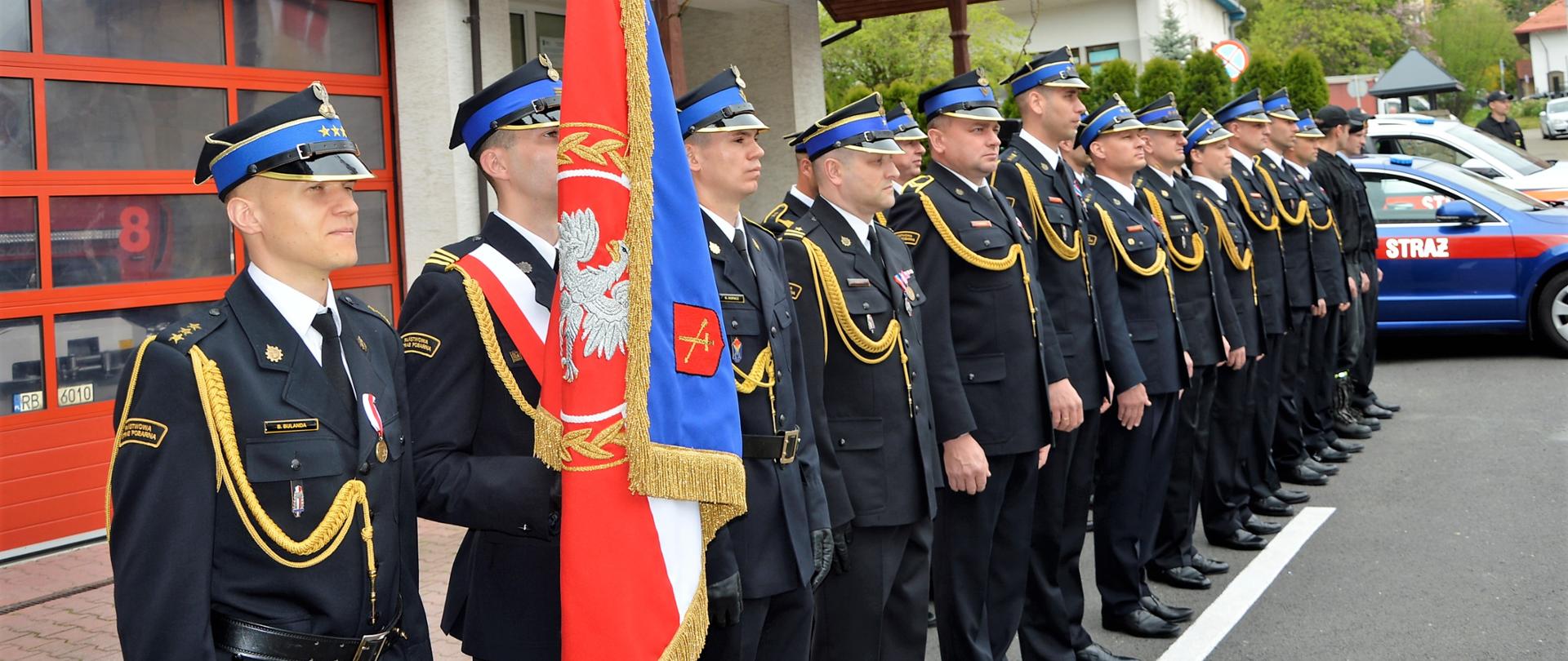 Zdjęcie przedstawia strażaków z KP PSP w Ustrzykach Dolnych ustawionych w dwuszeregu w mundurach galowych i służbowych na placu Komendy. Na pierwszym planie ustawiony jest poczet sztandarowy. W tle samochody Skoda superb i kodiak.