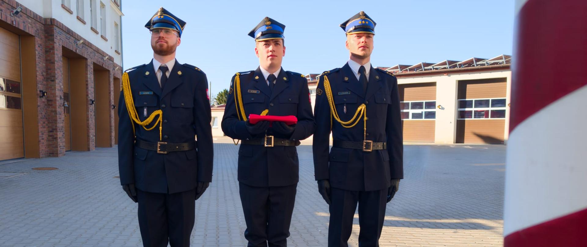 Na zdjęciu poczet flagowy:
kpt. Szczepan Zieliński
mł. kpt. Maciej Kuptz
sekc. Szymon Miler
Funkcjonariusze ubrani są w mundury galowe. sekc. Szymon Miler trzyma flagę Rzeczpospolitej Polskiej, która za moment zostanie uroczyście wciągnięta na maszt.