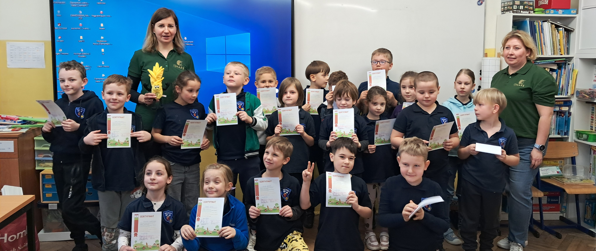 Zdjęcie grupowe dzieci po lekcji. Uczniowie stoją razem i uśmiechają się do zdjęcia, trzymając w rękach dyplomy. W tle widoczna tablica multimedialna w sali lekcyjnej