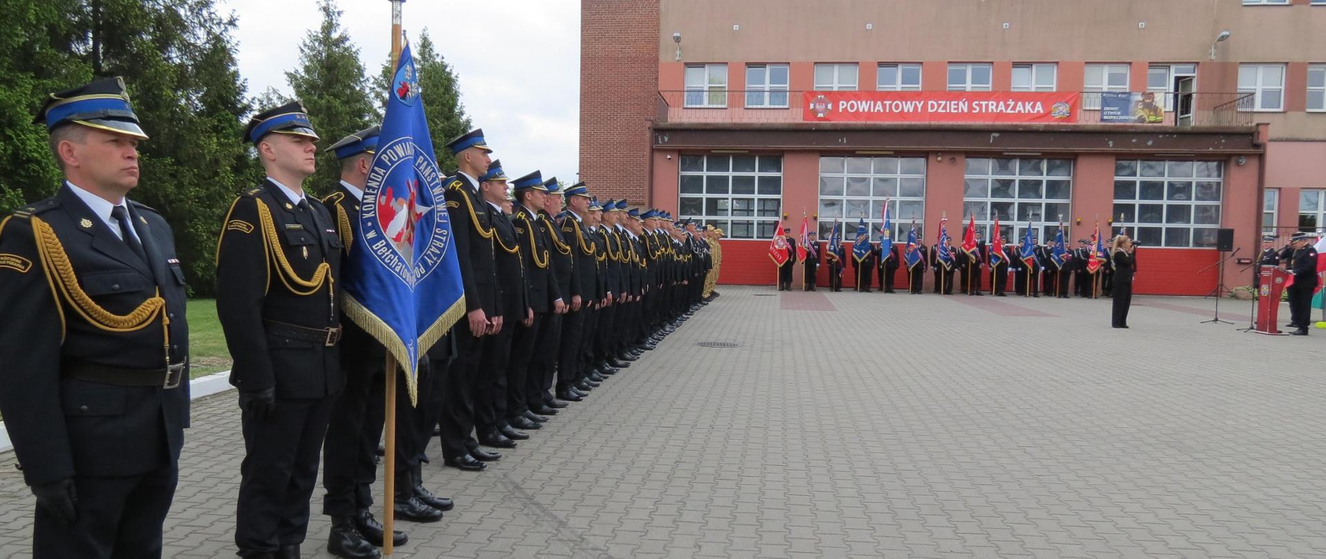 na zdjęciu widzimy strażaków w umundurowaniu wyjściowym w szyku w oddali budynek straży oraz poczet sztandarowy po prawo prowadzący ceremonię
