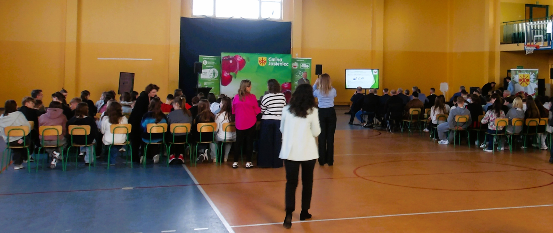 Grupa kilkudziesięciu osób siedzi na sali gimnastycznej przodem do ekranu, na którym prelegent prezentuje slajdy.