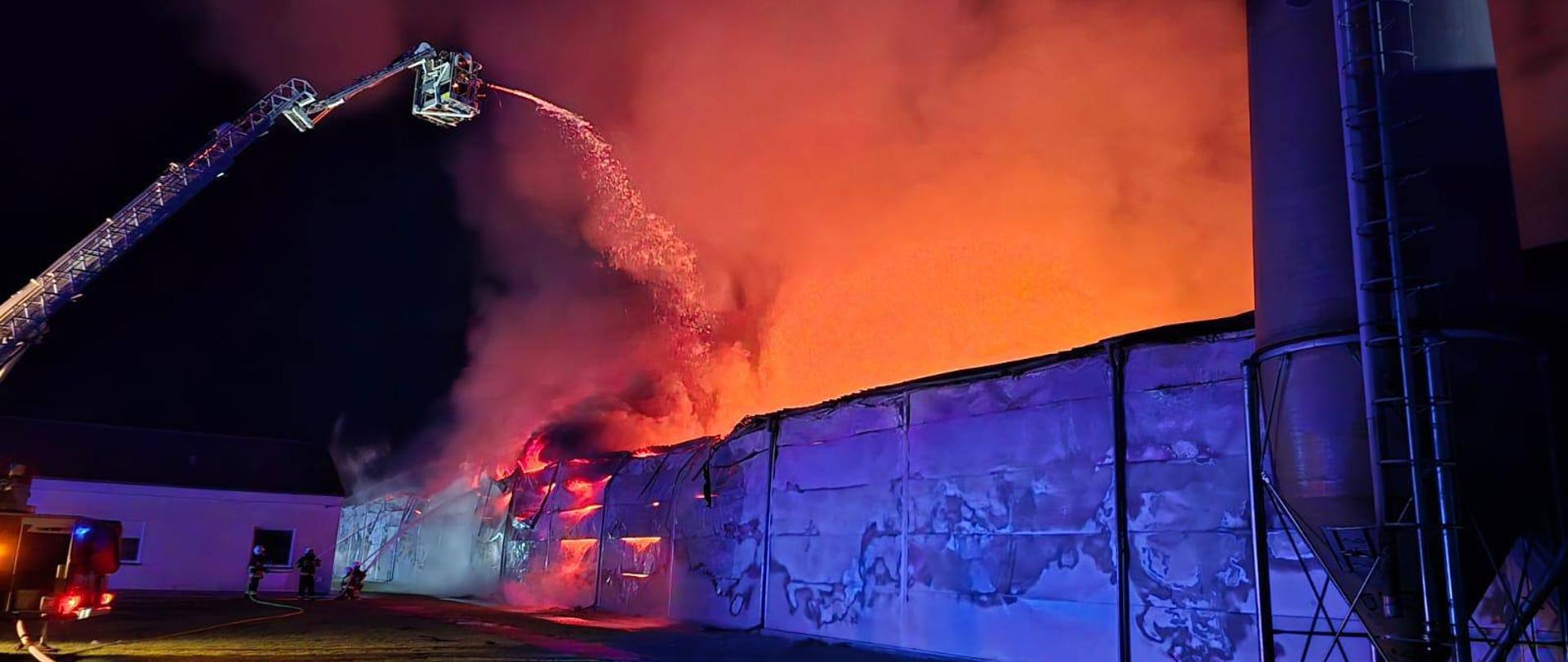 Duży budynek w ogniu nocą, z intensywnymi płomieniami i gęstym czarnym dymem unoszącym się z dachu. Strażacy gaszą pożar z wysięgnika wozu strażackiego, kierując strumień wody na płonącą konstrukcję. Budynek hali, a jego ściany są mocno uszkodzone. Scena oświetlona jest światłami awaryjnymi i blaskiem ognia.