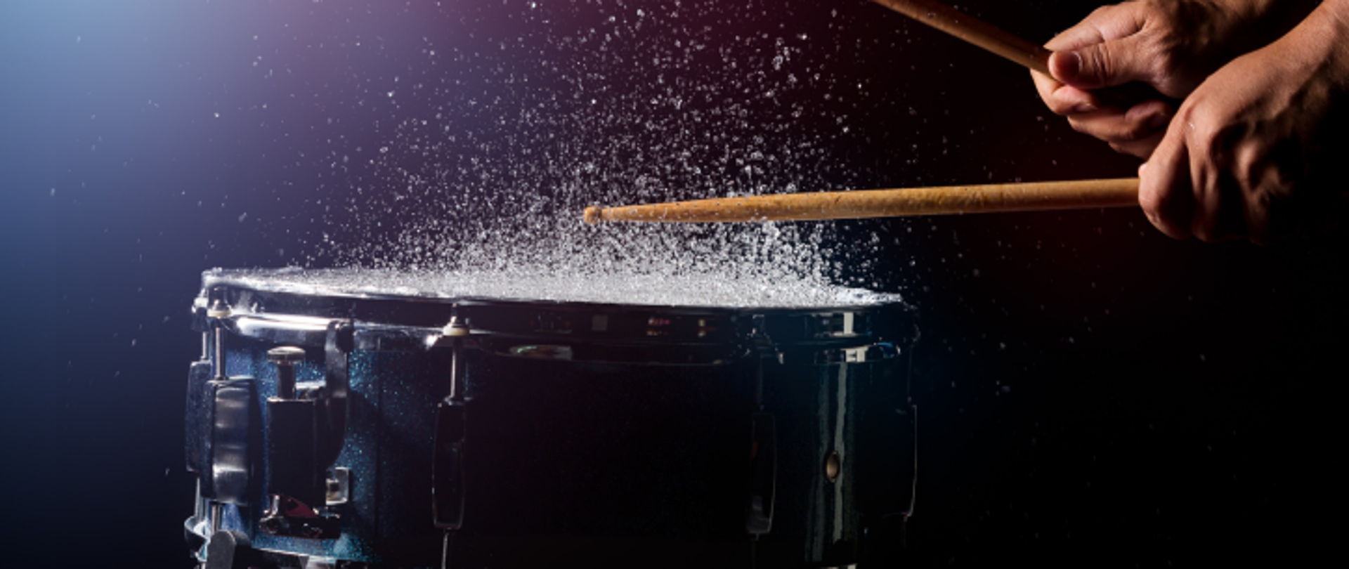 Granatowy bęben perkusyjny, z małą ilością wody na membranie, która przy sugerowanym na zdjęcu uderzeniu pałeczkami unosi się, tworząc deszczową mgłę nad bębnem