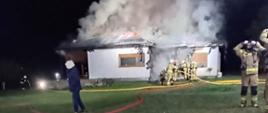 Zdjęcie przedstawia pożar budynku mieszkalnego jednorodzinnego. Nad domek widać silne zadymienie. Strażacy stoją przed budynkiem. Ponadto na zdjęciu widać linie wężowe.