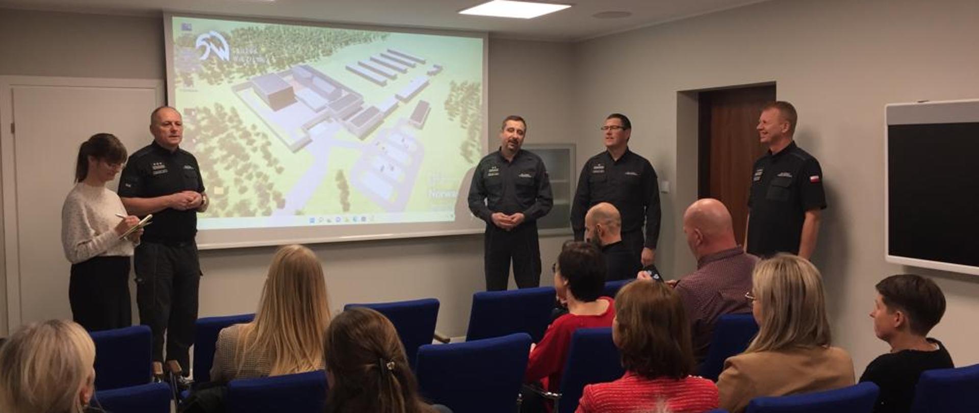 Prezentacja zakładu karnego w Kozichgłowach wyświetlana na projektorze na przedniej ścianie. Po bokach projektora osoby prezentująca, przedstawiciele Służby Więziennej. Widziana od tyłu publika na niebieskich krzesłach.