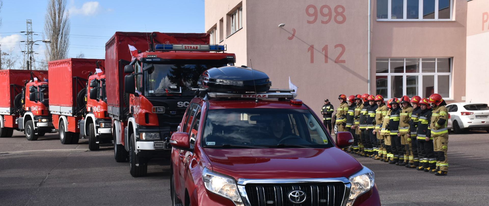 na zdjęciu kolumna czterech samochodów strażackich oraz dwunastu strażaków stojących na zbiórce w rzędzie w tle budynek z napisami 998 i 112