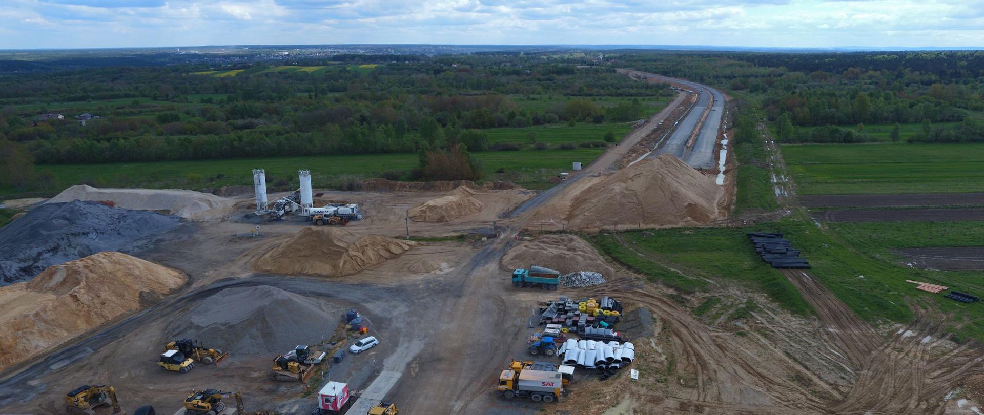 DK42 obwodnica Wąchocka - plac budowy sfotografowany z drona, maszyny, hałdy kruszywa i piasku, w oddali zarys dwujezdniowej trasy z warstwami bitumicznymi 