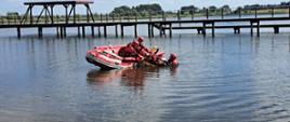 Zbliżenie na ratowników, którzy pracują na wodzie w czerwonej łodzi ratunkowej. W tle widać część pomostu, a cała scena odbywa się w słoneczny dzień. Dwóch ratowników znajdujących się w łodzi podejmuje na desce ratowniczej pozoranta z wody. Trzeci ratownik pracuje w wodzie dodatkowo asekurując pozorującą osobę.