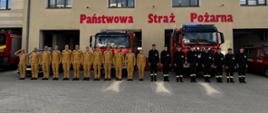 Plac manewrowy przed komenda w Rawiczu. Na tle budynku i stojących przed nią wozów bojowych, w szeregu ustawieni są strażacy. Ci stojący z lewej strony mają na sobie umundurowanie służbowe w kolorze piaskowym, ci z prawej - koszarowe w kolorze czarnym. Na budynku, nad bramami garazowymi widnieje czerwony napis PAŃSTWOWA STRAŻ POŻARNA