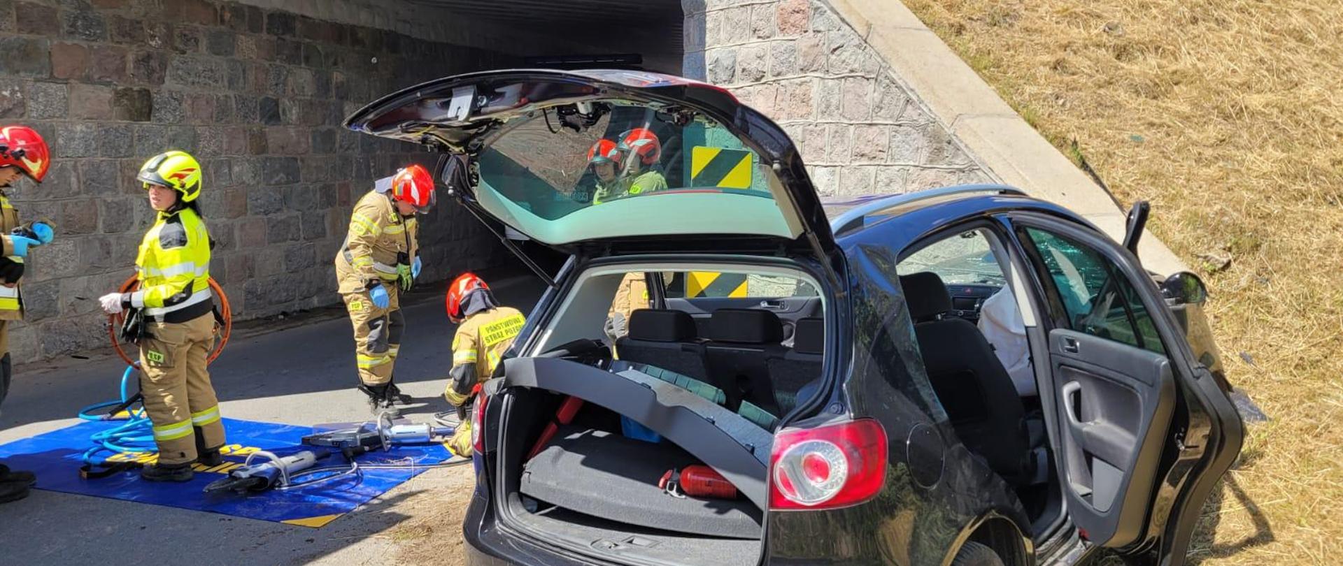 Słoneczny dzień na tle szarego betonowo - kamiennego wiaduktu kolejowego widoczny jest tył czarnego samochodu osobowego, który przodem uderzył w wiadukt. Klapa samochodu i prawe tylne drzwi są otwarte. Z lewej strony widać strażaków pięciu strażaków ubranych w piaskowe ubrania specjalne i czerwone hełmy. Szósty strażak ubrany jest w żółtą odblaskową kurtkę przeznaczoną do ratownictwa technicznego i żółty hełm. Na wiadukcie widać nogi pracowników kolei ubranych w pomarańczowe uniformy. W prawym górnym rogu widać żółtą koparkę. Trawa w miejscu zdarzenia ma kolor blado-zielony(siana).