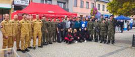 Zdjęcie przedstawia dużą grupę osób, w tym: strażaków, żołnierzy i przedstawicieli innych służb pozujących wspólnie do fotografii. W tle znajdują się namioty oraz zabudowania miejskiego rynku. Uczestnicy są ubrani w mundury służbowe.