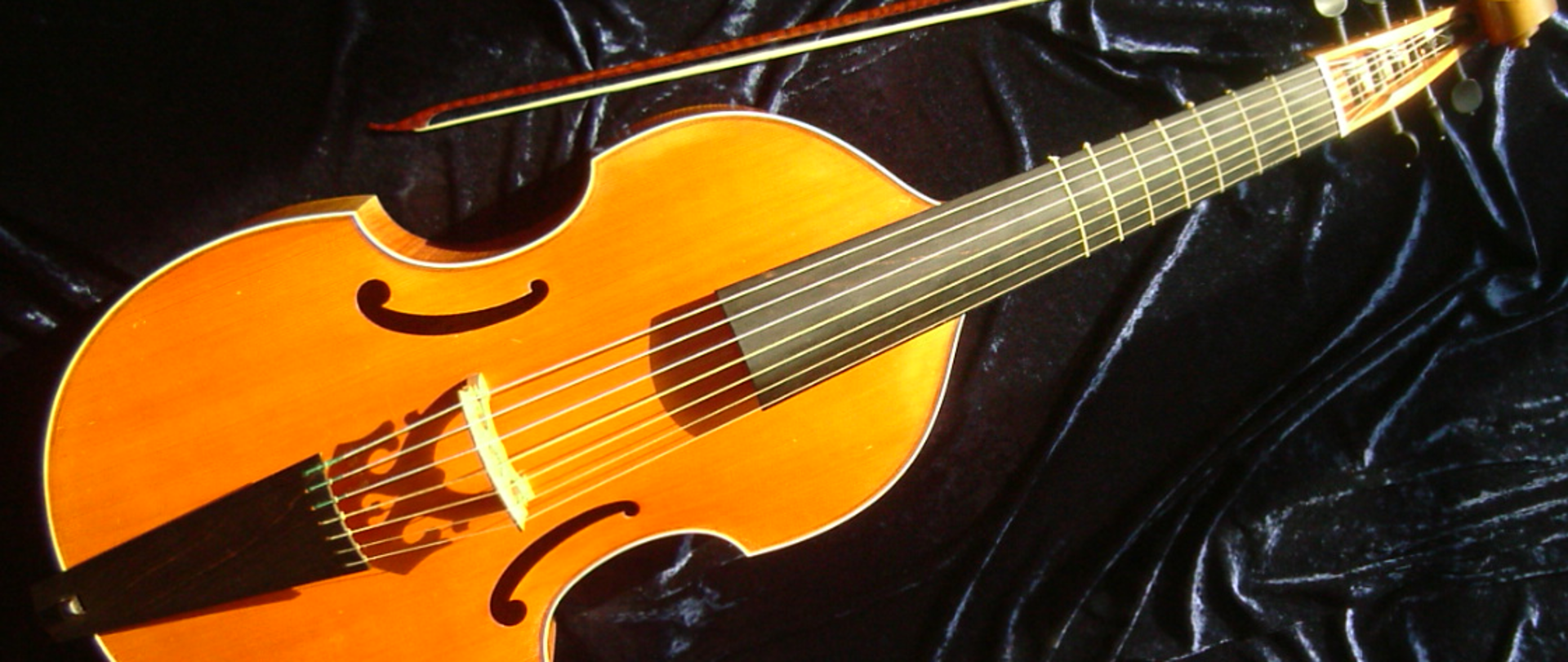 Zdjęcie przedstawia instrument Viola da gamba na czarnym tle. Obok instrumentu leży smyczek.