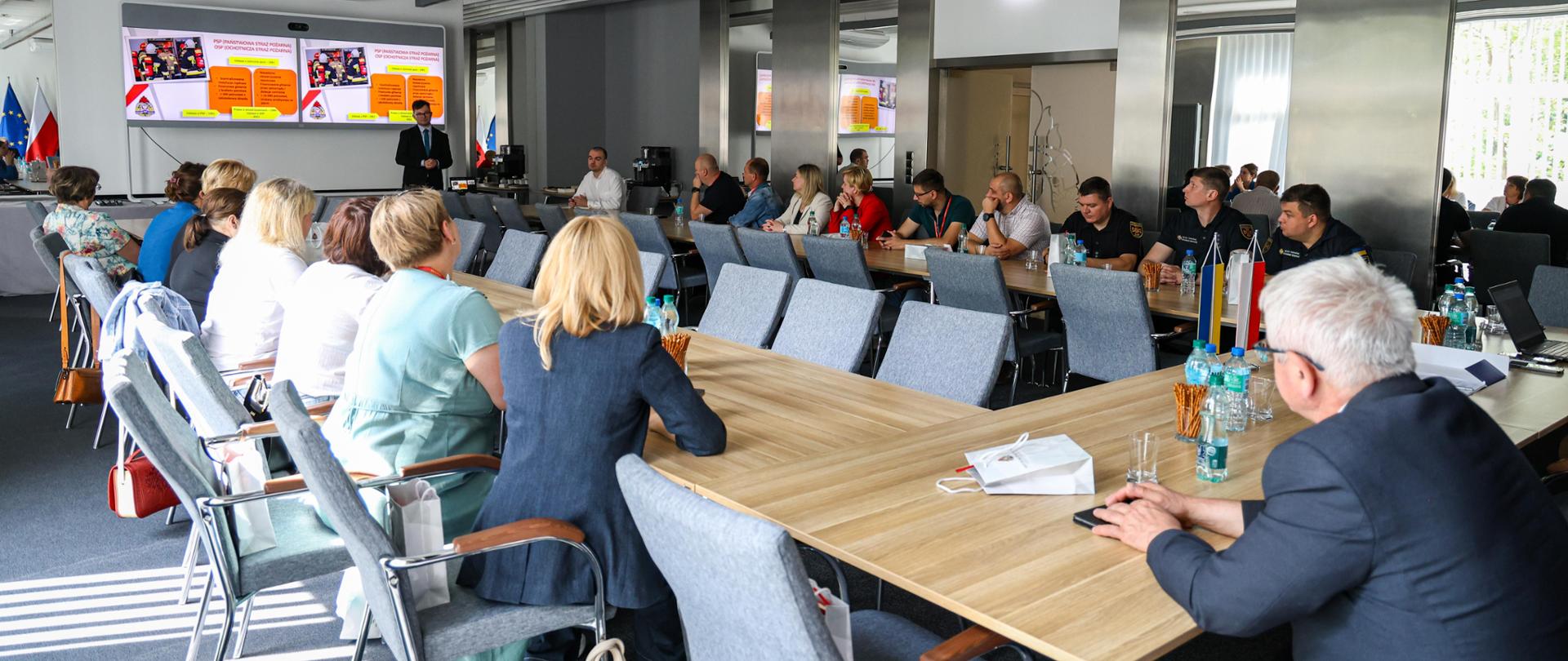 Uczestnicy spotkania siedzą w sali. mężczyzna prowadzi prelekcję