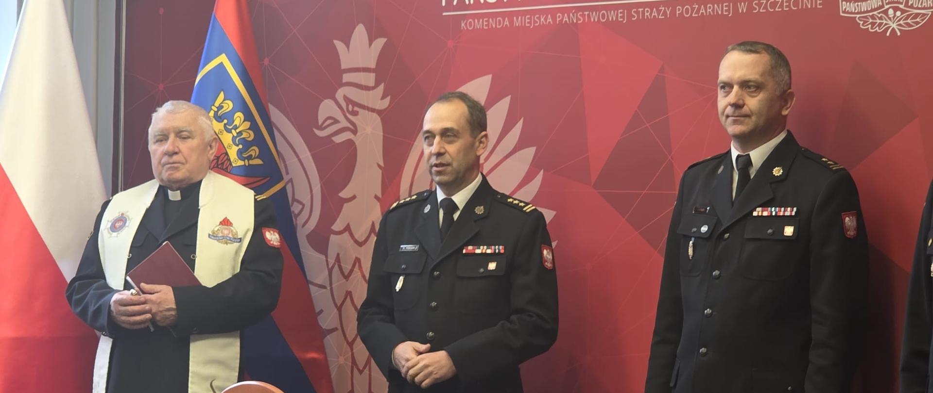 Na fotografii widoczne są trzy postacie stojące przed czerwoną ścianką z logotypem Państwowej Straży Pożarnej:
Po lewej stronie znajduje się duchowny, prawdopodobnie kapelan straży pożarnej, ubrany w sutannę i biało-złotą stułę z motywami religijnymi (m.in. wizerunkiem św. Floriana, patrona strażaków).
W centrum i po prawej stronie stoją dwaj oficerowie Państwowej Straży Pożarnej w mundurach wyjściowych. Wielkanoc 2026