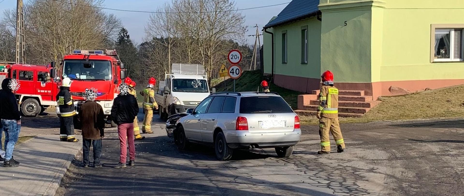 Rozbite samochody osobowe, pracujący strażacy i osoby poszkodowane. Z lewej strony 2 samochody pożarnicze