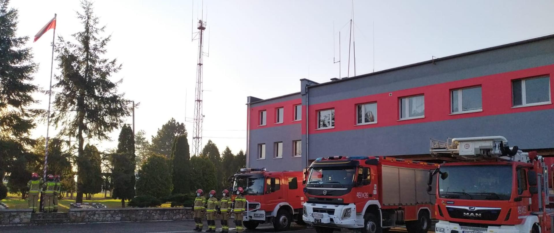 Na zdjęciu znajdują się strażacy, którzy podnoszą flagę państwową, samochody pożarnicze na tle budynku komendy