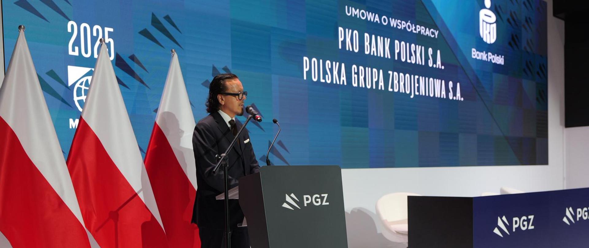 Minister Wojciech Balczun stoi za mównicą. W tle ekran z napisem PKO Bank Polski S.A., Polska Grupa Zbrojeniowa S.A. i flagi biało-czerwone.