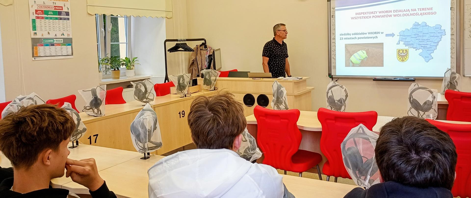 W sali lekcyjnej uczniowie siedzą przy ławkach wyposażonych w osłonięte słuchawki nauszne. Na pierwszym planie widać trzech uczniów obserwujących prezentację prowadzoną przez inspektora. Na ekranie wyświetlana jest mapa województwa dolnośląskiego oraz informacje dotyczące siedzib Oddziałów Inspekcji w województwie dolnośląskim.
