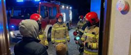 Na zdjęciu widoczni są strażacy Państwowej Straży Pożarnej prowadzący działania kontrolne przy wejściu do budynku mieszkalnego w porze nocnej. Jeden z ratowników obsługuje przenośny detektor do pomiaru stężenia niebezpiecznych gazów, a pozostali pozostają w gotowości operacyjnej. W tle znajduje się samochód ratowniczo-gaśniczy z włączonym oświetleniem ostrzegawczym, oświetlający miejsce zdarzenia. Przy wejściu do budynku widoczna jest czujka tlenku węgla, która sygnalizuje potencjalne zagrożenie. Cała scena obrazuje profesjonalne i spokojne działania strażaków mające na celu zapewnienie bezpieczeństwa mieszkańcom.