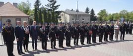 Na zdjęciu widzimy strażaków oraz osoby cywilne stojące w szeregu. Strażacy ubrani są w galowe mundury w ręku trzymają teczki koloru czerwonego. W tle widzimy budynki mieszkalne, zielone drzewa 