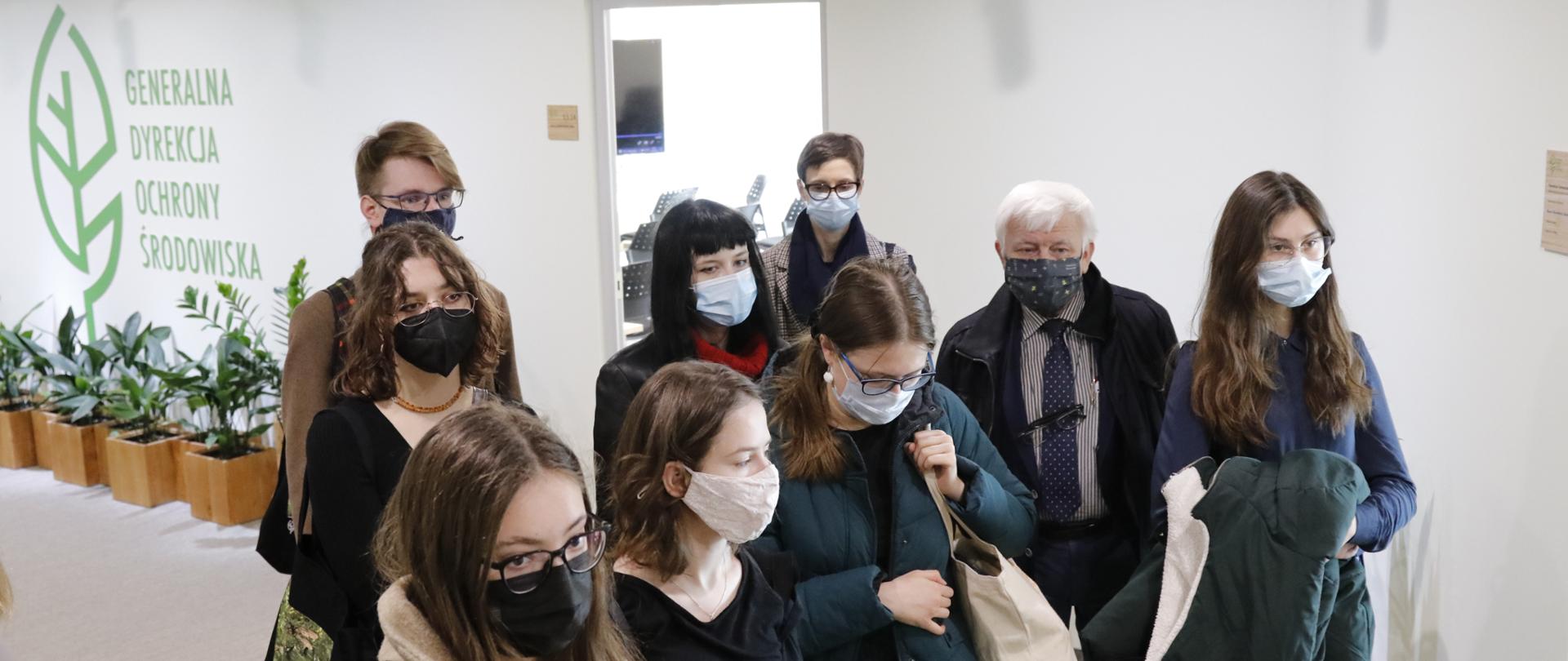 Korytarz. Na tle ściany z logo Generalnej Dyrekcji Ochrony Środowiska (białe tło, na nich logotypy - zielony liść z napisem Generalna Dyrekcja Ochrony Środowiska) stoi grupa młodych osób, kobiety i mężczyźni. W centralnej części jeden widać dojrzałego mężczyznę o siwych włosach. 